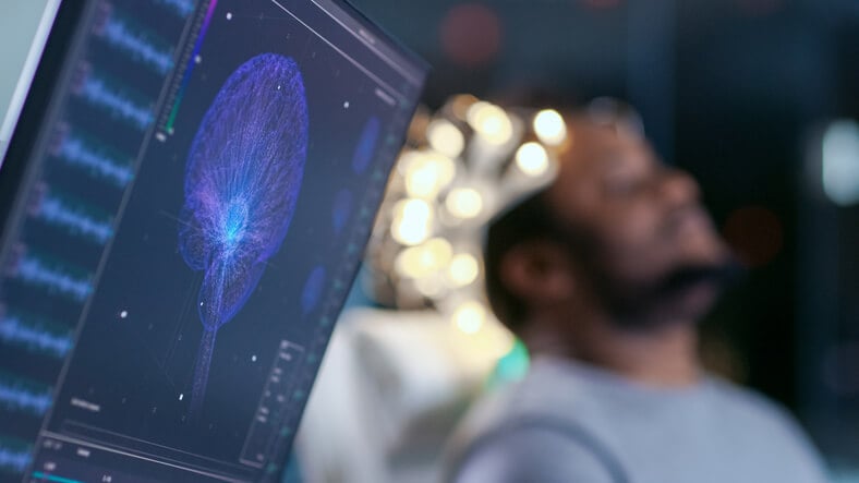 brain scan machine in foreground with patient in background in dark room with lit nodes attached to skull