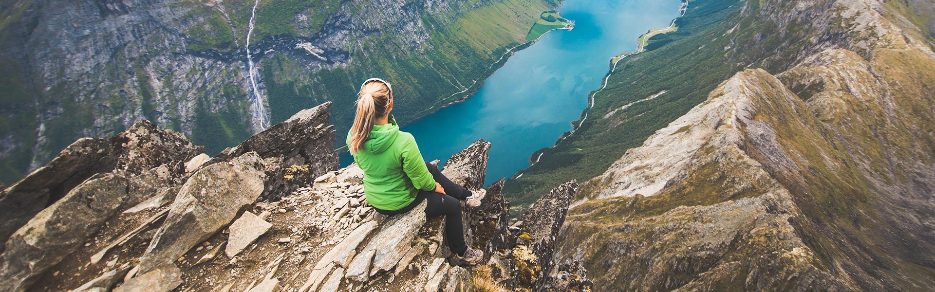 Female nurse in green jacket sitting on mountain top looking down into water.