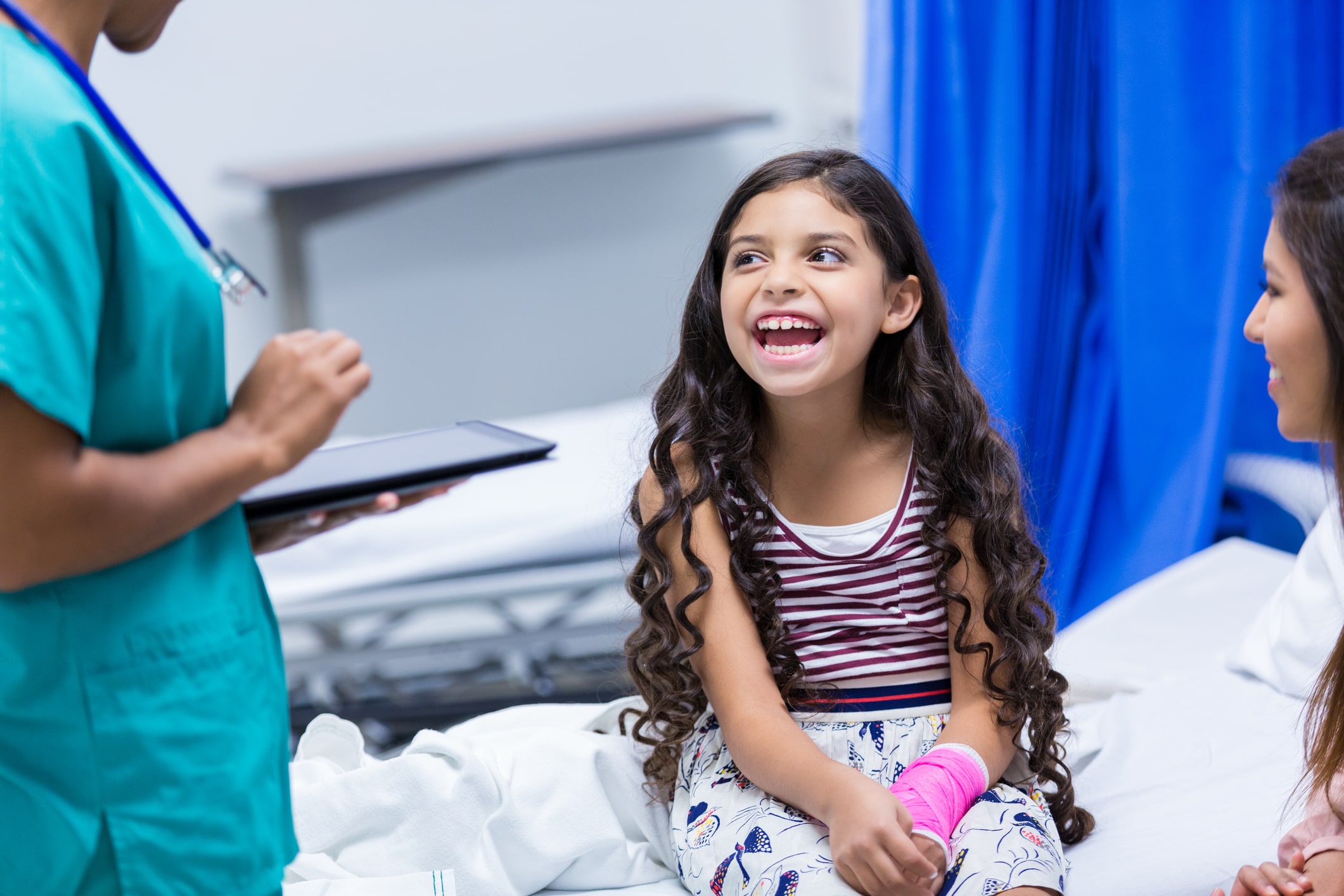 nurse in green scrubs holding ipad, standing over young white brunette girl smiling sitting on hospital bed nurse in green scrubs holding ipad, standing over young white brunette girl smiling sitting on hospital bed