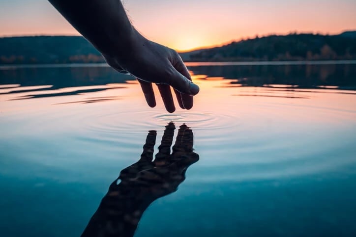 persons hand touching water with setting sun behind