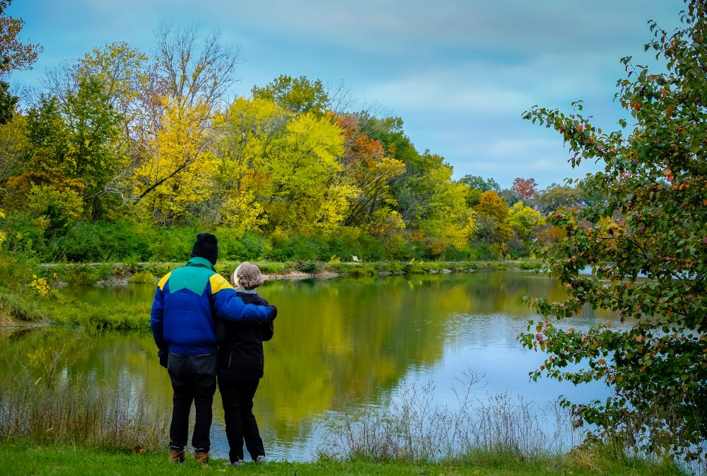 Couple hugging standing near a lake in a wooded area.