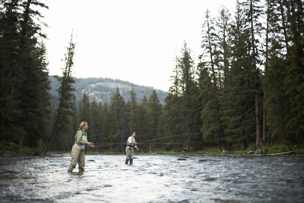 Two people standing in water wearing fishing gear and holding poles in the water.